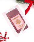 A pink novelty ornament bar shaped like a US passport, displayed in a clear windowed pink gift box. The box is photographed on a white background with holiday props, including red berries and a red ribbon.