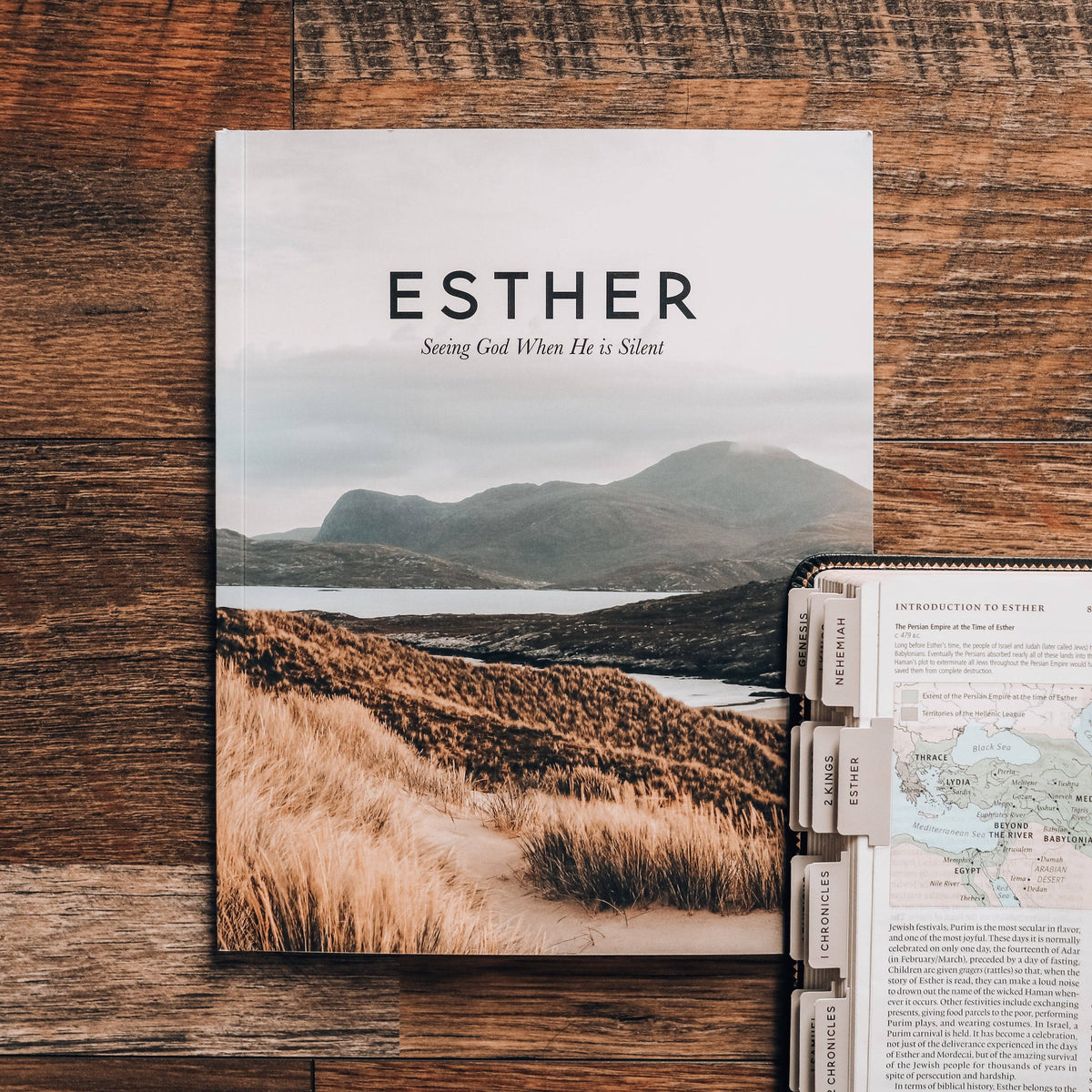 A book titled 'Esther' with a serene mountainous landscape on the cover, positioned next to a map and other books on a wooden surface.