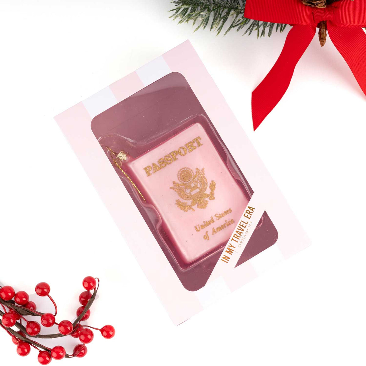 A pink novelty ornament bar shaped like a US passport, displayed in a clear windowed pink gift box. The box is photographed on a white background with holiday props, including red berries and a red ribbon.