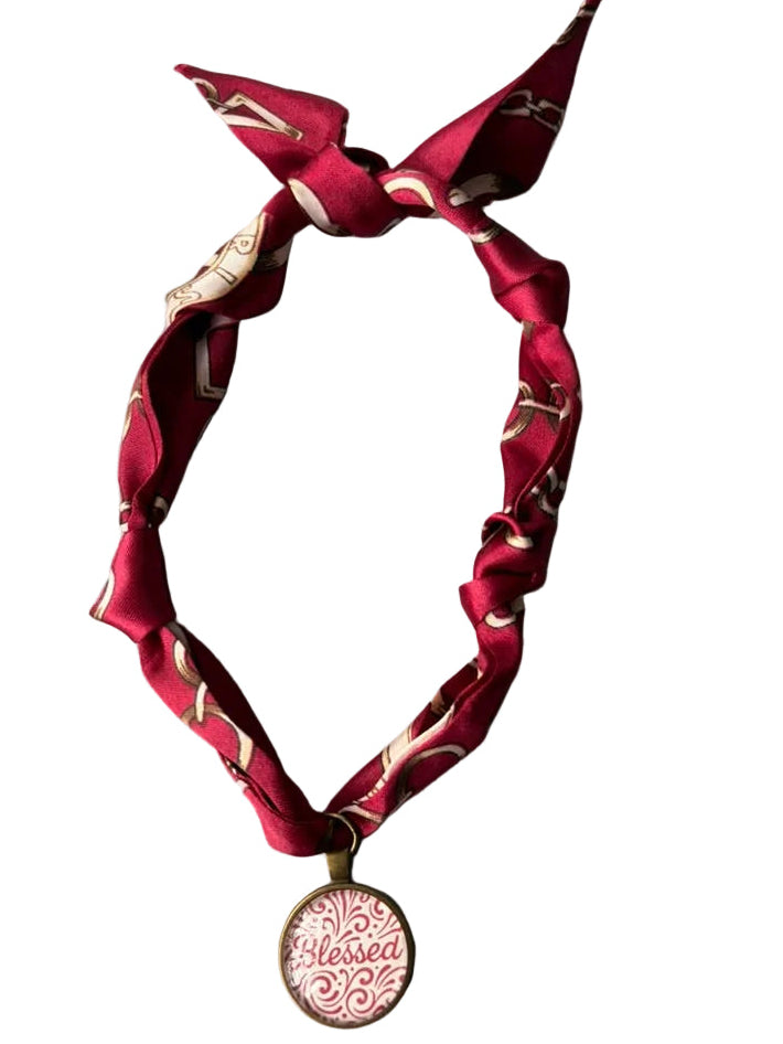 A red satin neck scarf with a gold pattern, tied in a knot at the top and hanging from a gold chain with a round pendant that reads 'Blessed'.