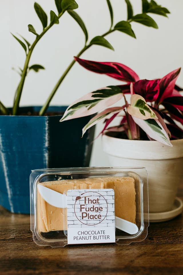 A plastic-wrapped square of chocolate peanut butter fudge on a wooden surface with a plant in the background.
