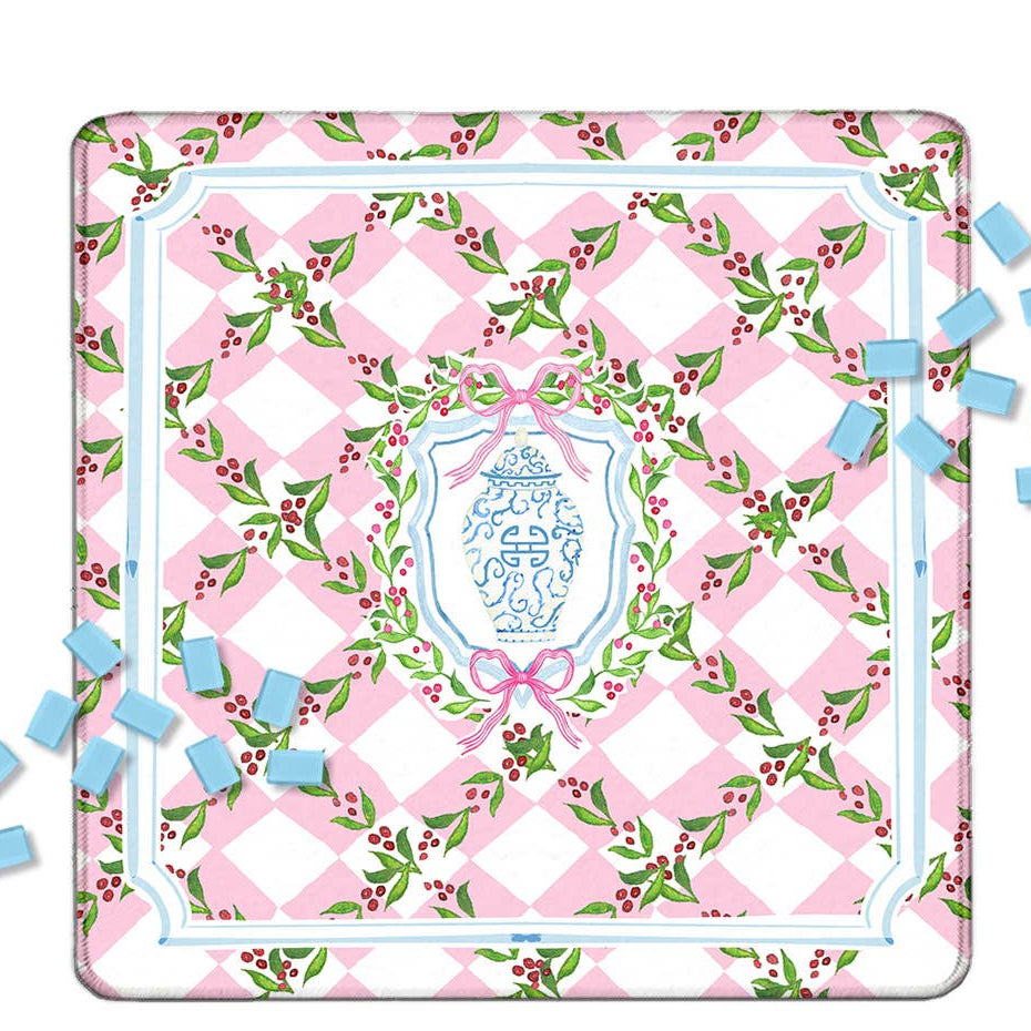 A square decorative Mahjong mat with a pink and white diamond pattern, green holly leaves, and red berries, featuring a central blue and white crest surrounded by a pink bow.  Light blue Mahjong tiles are scattered around it on a white background.