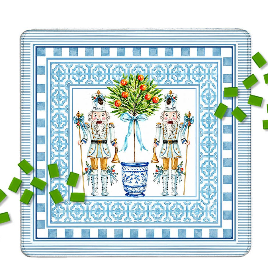A square decorative mahjong mat featuring two nutcracker figures flanking a blue floral pot with red berries, surrounded by ornate blue patterned borders. Green mahjong tiles are scattered around the edges on a white background.