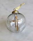 A clear glass Christmas ornament with a gold metal cap, gold hanging ribbon, and a wooden cross inside, set on a light marble surface.
