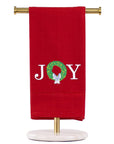 A red kitchen towel hanging from a gold stand with a white marble base, featuring the word 'JOY' embroidered in white with a green wreath and white bow.
