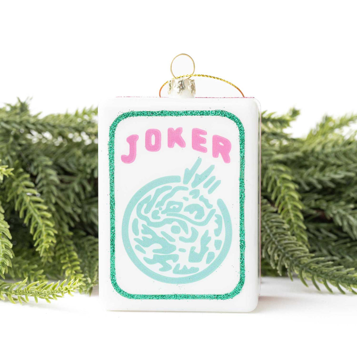 Christmas ornament with 'Joker' text and design, hanging against a white background with greenery.