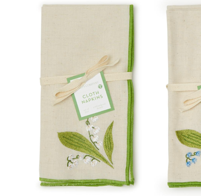 Two folded cloth napkins on a white background. Each napkin is made of natural linen with a green border, decorated with embroidered botanical motifs, and tied with a ribbon and paper tag that reads CLOTH NAPKINS.