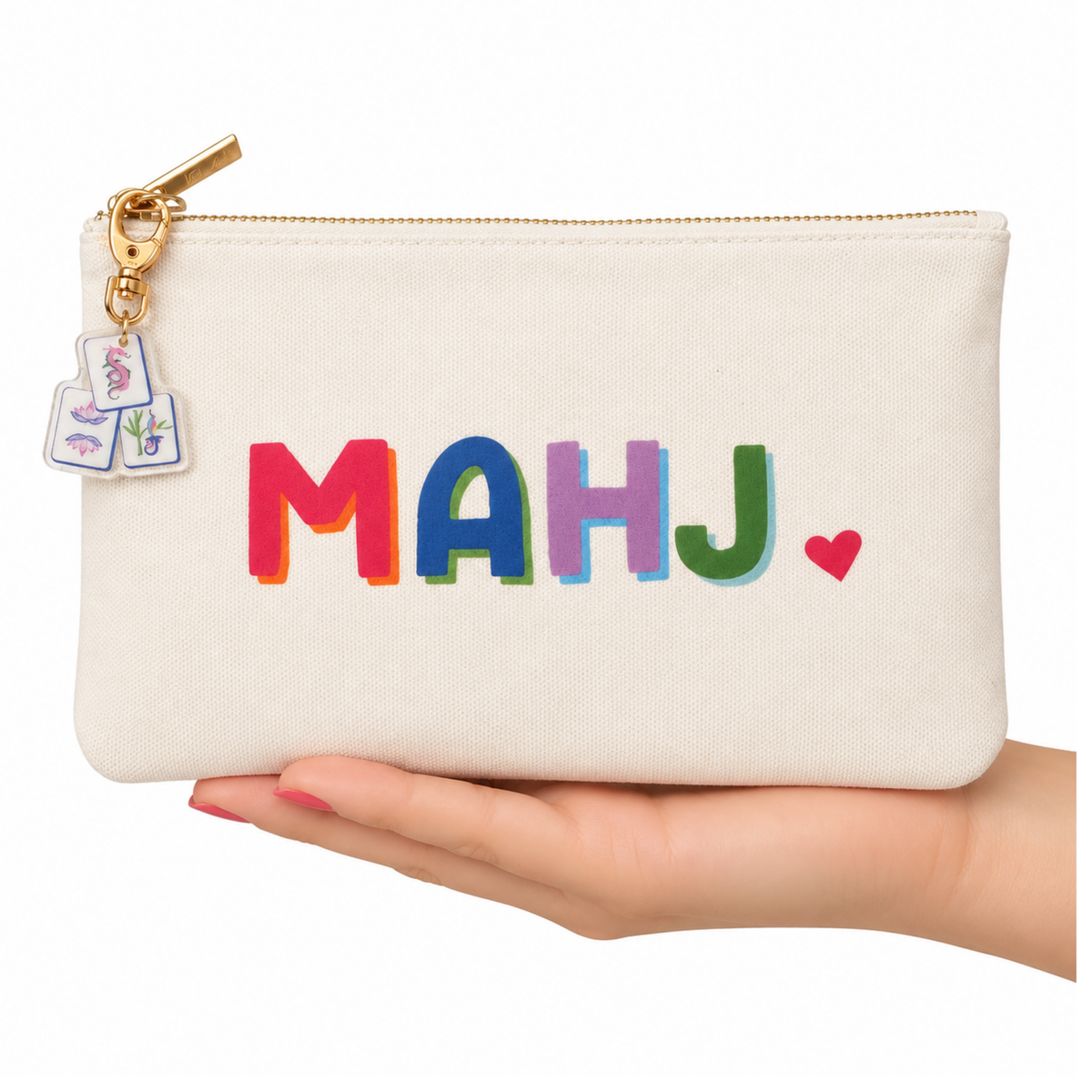 A cream canvas zip pouch with the word 'MAHJ' in colorful embroidered letters and a small red heart, held in a hand. The pouch has a gold zipper and a small embroidered key charm.