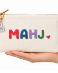 A cream canvas zip pouch with the word 'MAHJ' in colorful embroidered letters and a small red heart, held in a hand. The pouch has a gold zipper and a small embroidered key charm.