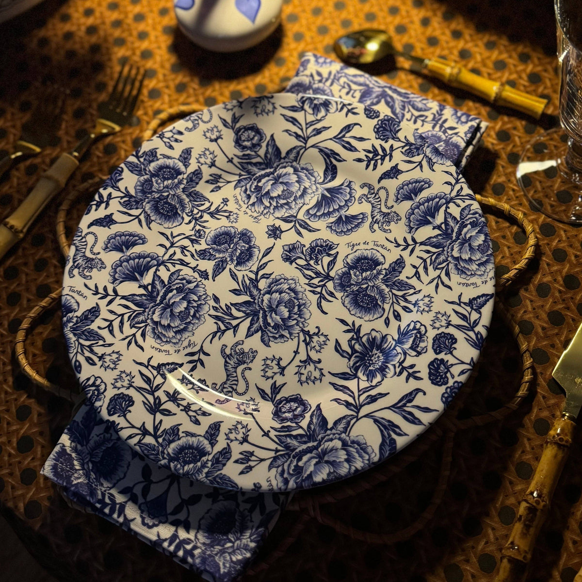 A blue and white floral patterned melamine dinner plate on a table setting with cutlery and a orange patterned tablecloth.