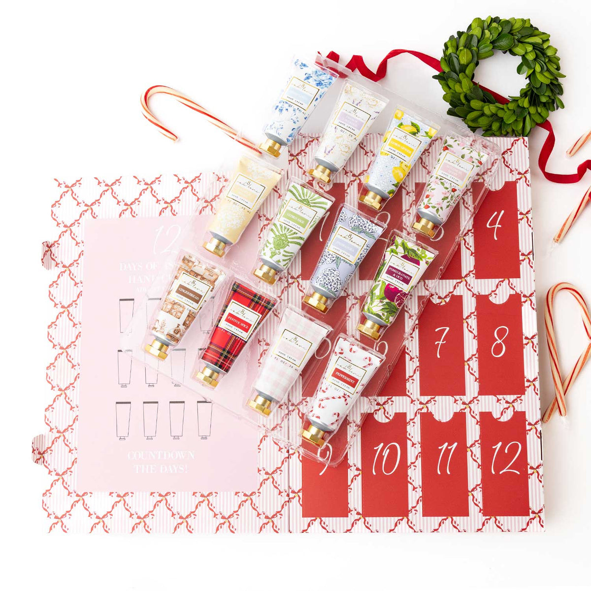 A Christmas advent gift set featuring 12 hand cream tubes arranged in a clear plastic tray over a pink and red advent calendar, with candy canes, a green wreath, and ribbon accents on a white background.