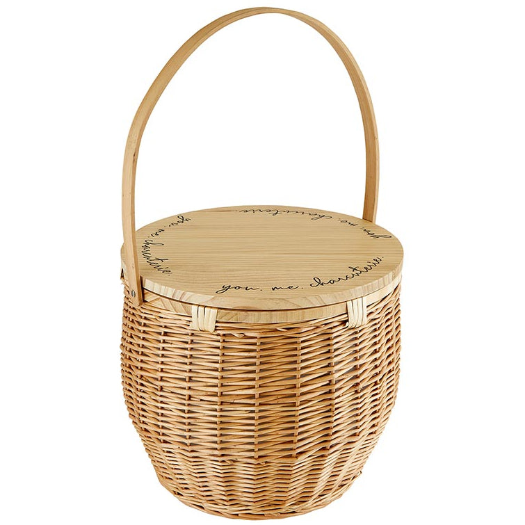 A beige picnic basket with a wicker body and a wooden lid, which has the words 'you & me. charcuterie' engraved on it.