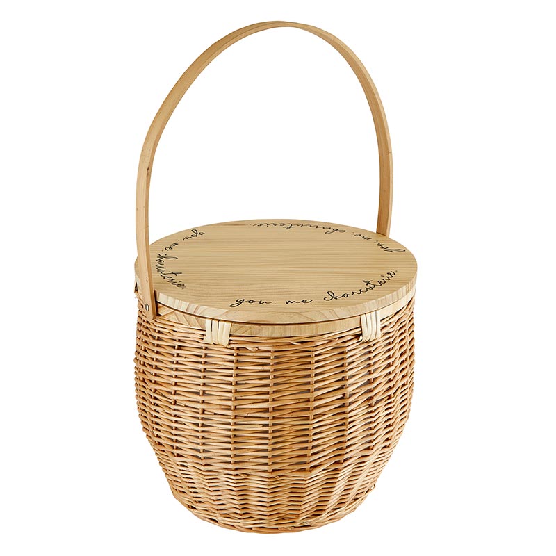 A beige picnic basket with a wicker body and a wooden lid, which has the words 'you & me. charcuterie' engraved on it.