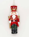 A classic Christmas nutcracker ornament hanging against a plain white background. The ornament features a red coat with gold trim, a red hat with gold accents, a white beard, and green and black pants.