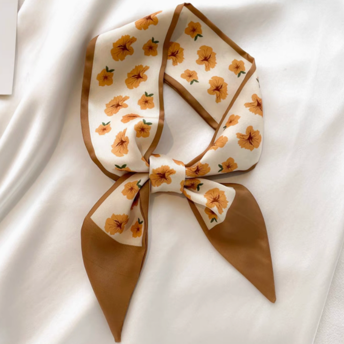 A cream-colored silk scarf with a brown border and a pattern of orange flowers, tied in a bow and laid flat on a white fabric background.