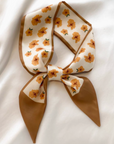 A cream-colored silk scarf with a brown border and a pattern of orange flowers, tied in a bow and laid flat on a white fabric background.