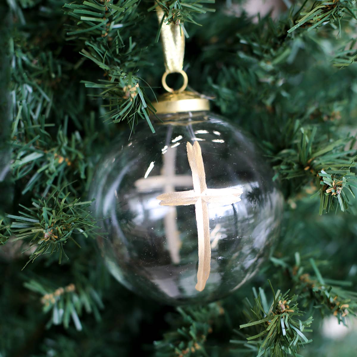A clear glass Christmas ornament hanging on a green Christmas tree, featuring a light wood cross printed inside and a gold hanging loop at the top.